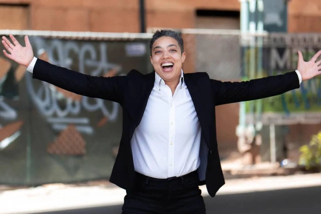 Chelsea Downs founder of New York Toy Collective. Arms open in a white shirt with black blazer.